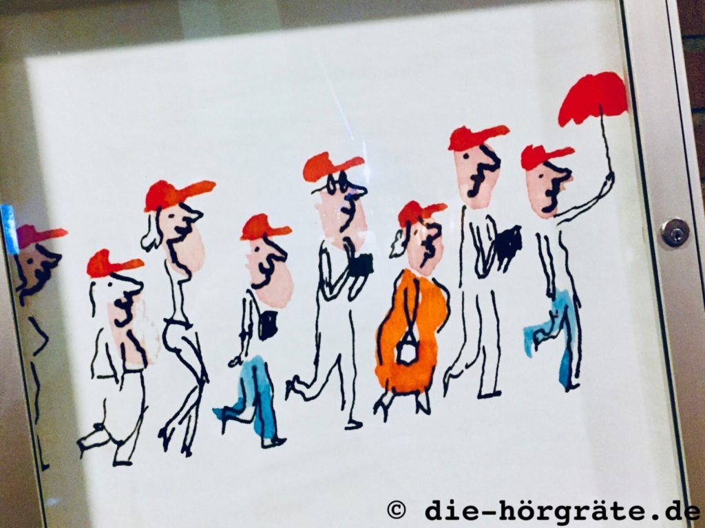 Zeichnung von Leuten, die hintereinander laufen und alle rote Schirmmützen aufhaben, die Zeichnung hänt in einem Glaskasten