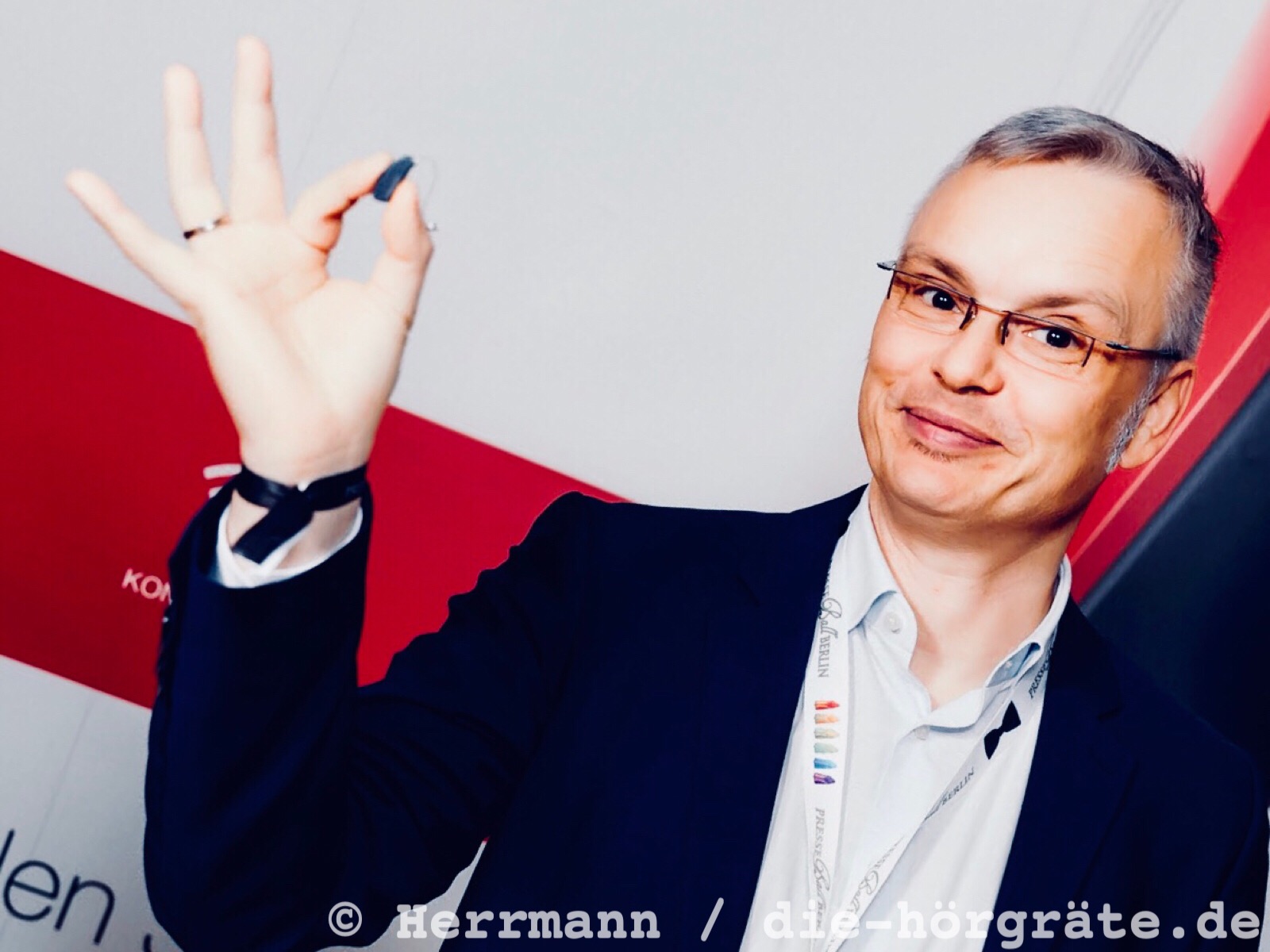 der Blog-Autor Martin Schaarschmidt lächelt in die Kamera und zeigt ein kleines Hörgerät, das er zwischen Daumen und Zeigefinger hält